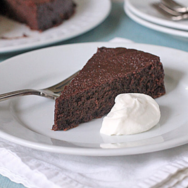 A slice of rich chocolate cake on a white plate with a dollop of whipped cream, accompanied by a fork. The plate rests on a white napkin, and other plates and forks are visible in the background.