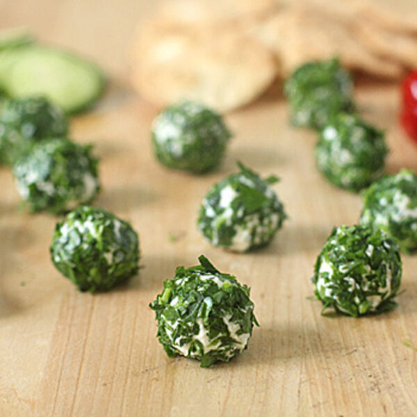 Small cheese balls coated in chopped herbs are arranged on a wooden surface, with sliced cucumber, black olives, red peppers, and pita pieces in the background.
