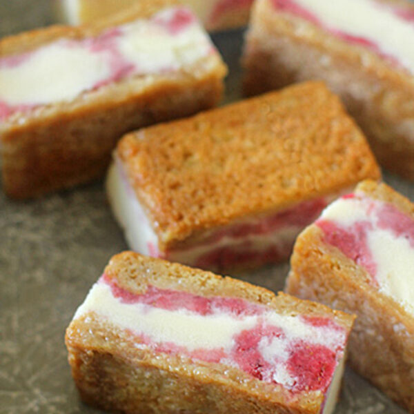 Rectangular ice cream sandwiches with golden-brown cookie edges and a layer of vanilla ice cream swirled with pink fruit filling, arranged on a gray textured surface.