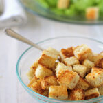 A glass bowl filled with golden, toasted croutons sits on a light wooden table, with a spoon inside and a blurred plate of green salad in the background.
