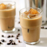 Two glasses of iced coffee with milk, each filled with ice cubes, sit on a white surface. Coffee beans are scattered around the glasses, and a metal pitcher is visible in the background.