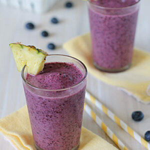Two glasses of purple blueberry smoothie, each garnished with a pineapple wedge, sit on yellow napkins. Striped straws and scattered blueberries are nearby, with a white container of blueberries in the background.