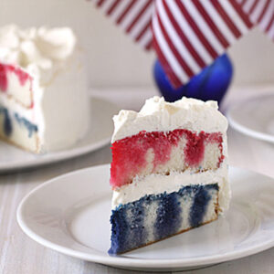 A slice of layered cake with red, white, and blue sections and white frosting sits on a plate. An American flag and another slice of cake are visible in the background.