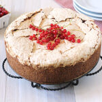 A round cake with a cracked, golden-brown top is displayed on a metal stand, decorated with a bunch of red currants. A small basket of red currants and a stack of plates are in the background.