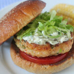 A chicken burger with lettuce, tomato, and creamy sauce on a toasted bun, served on a white plate with a side of corn on the cob.