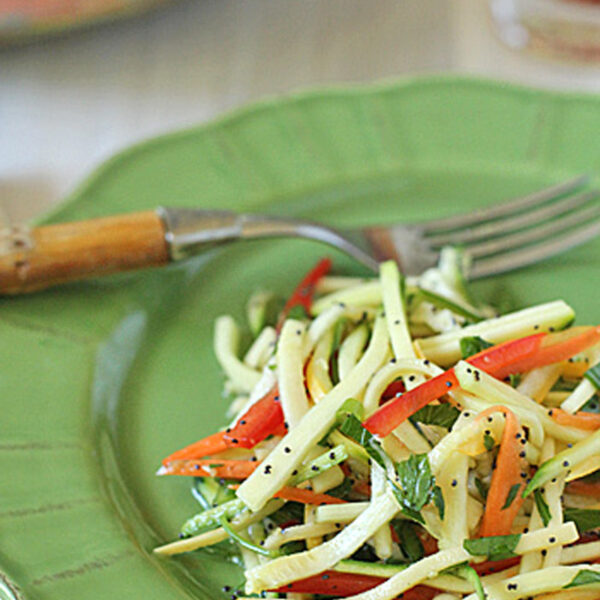 A green plate holds a fresh salad made of thinly sliced zucchini and red bell peppers, garnished with herbs and poppy seeds. A fork rests on the plate.
