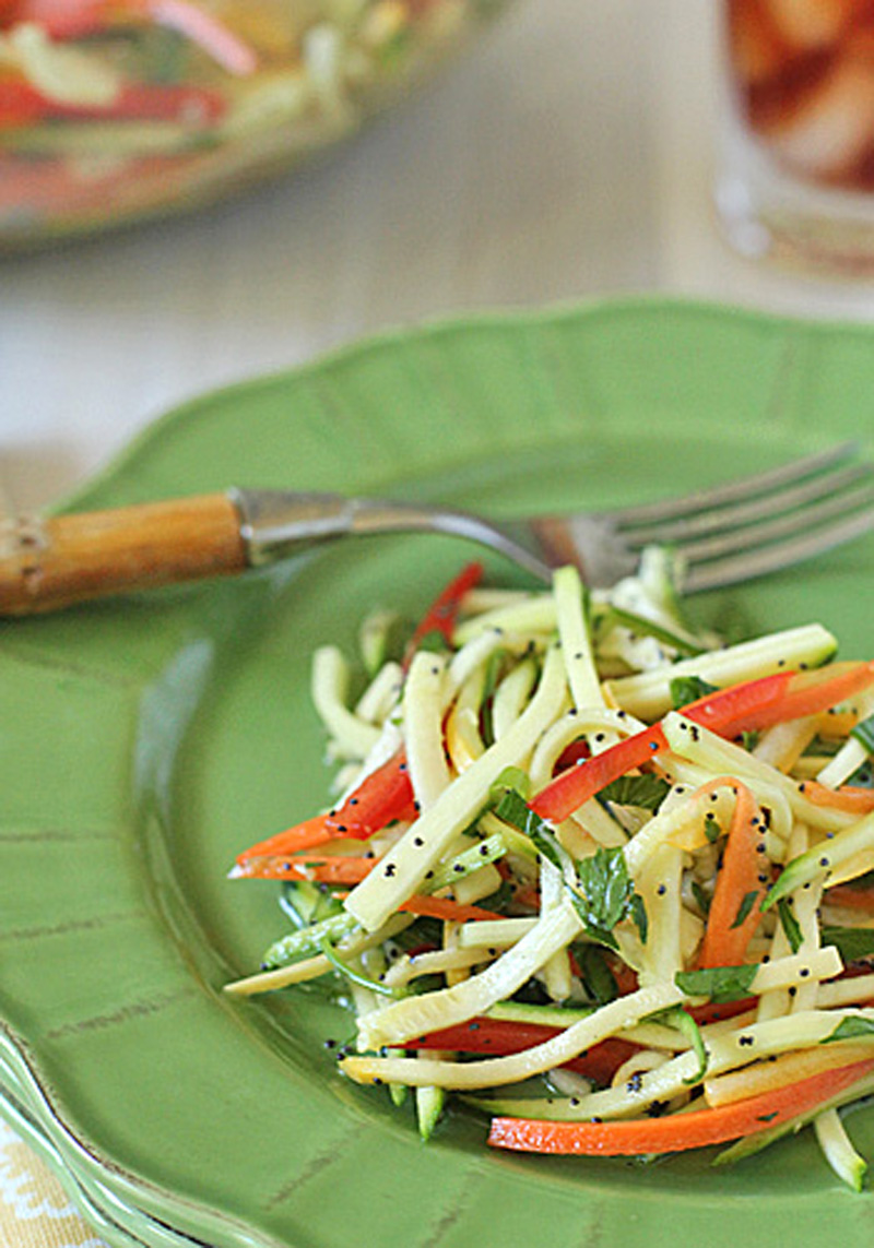 A green plate holds a fresh salad made of thinly sliced zucchini and red bell peppers, garnished with herbs and poppy seeds. A fork rests on the plate.