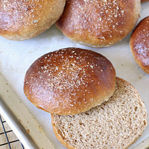 Several whole wheat sandwich buns on a baking sheet lined with parchment paper, with one bun sliced open to show the soft, fluffy interior. A cooling rack is partially visible beside the tray.