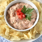 A bowl of creamy dip topped with chopped tomatoes and parsley, surrounded by potato chips on a white plate, set on a light blue napkin.