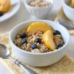 A white bowl of oatmeal topped with peach slices, blueberries, and slivered almonds sits on a yellow patterned napkin, with a spoon beside it. Another bowl and plate with similar ingredients are in the background.