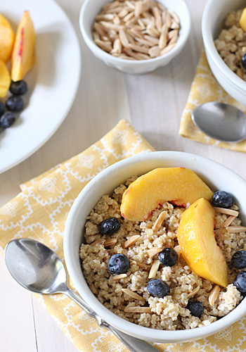 A bowl of cooked cereal topped with fresh peach slices, blueberries, and slivered almonds, set on a yellow patterned napkin. A spoon is beside the bowl, and there are additional bowls and toppings in the background.