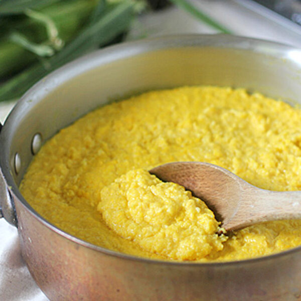 A wooden spoon stirs a pot of creamy yellow polenta in a metal saucepan, with fresh corn partially visible in the background.