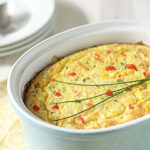 A baked corn casserole garnished with chives sits in a light blue ceramic dish on a yellow patterned napkin, with plates and spoons in the background.