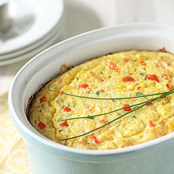 A baked corn casserole garnished with chives sits in a light blue ceramic dish on a yellow patterned napkin, with plates and spoons in the background.