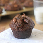 A chocolate muffin with chocolate chips sits on a white napkin, with a glass of milk and a muffin tray with more muffins in the blurred background.