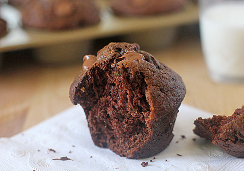 A chocolate muffin with a bite taken out, sitting on a white napkin. Melted chocolate chips are visible on top, and a glass of milk and muffin tray are blurred in the background.