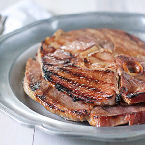 Slices of grilled ham steak stacked on a silver plate, with a fork and knife placed on a white napkin in the background.