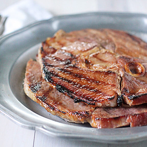 Slices of grilled ham steak stacked on a silver plate, with a fork and knife placed on a white napkin in the background.