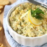 A white dish filled with creamy artichoke dip, garnished with a lemon slice and parsley, sits on a blue cloth. Crackers are arranged nearby on a wooden surface, ready for dipping.