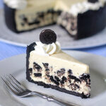 A slice of cookies and cream cheesecake with a chocolate crust and whipped cream, topped with a mini chocolate cookie, sits on a plate with a fork. The rest of the cheesecake is in the background on a cake platter.