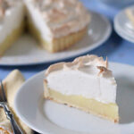 A slice of lemon meringue pie with a golden crust, yellow lemon filling, and fluffy meringue topping sits on a white plate, with the rest of the pie and a fork visible in the background.