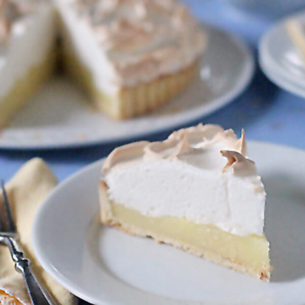 A slice of lemon meringue pie with a golden crust, yellow lemon filling, and fluffy meringue topping sits on a white plate, with the rest of the pie and a fork visible in the background.