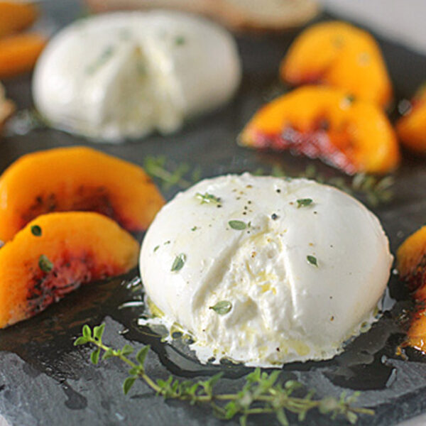 A slate platter with two balls of burrata cheese, sliced yellow peaches, sprigs of thyme, toasted bread slices, and a drizzle of olive oil.