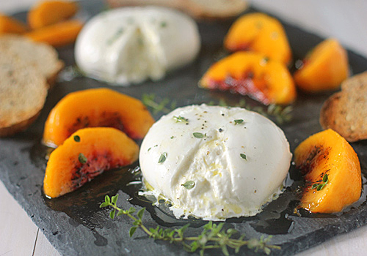 A slate platter with two balls of burrata cheese, sliced yellow peaches, sprigs of thyme, toasted bread slices, and a drizzle of olive oil.