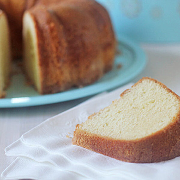 A single slice of plain bundt cake sits on a white napkin, with the rest of the cake in the background on a light blue plate. The cake has a golden brown crust and a light, fluffy interior.