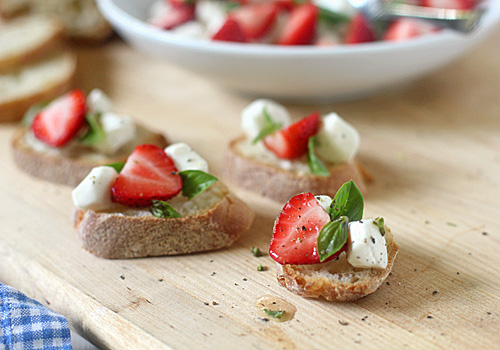 Slices of baguette topped with fresh mozzarella, strawberry halves, green basil leaves, and a sprinkle of black pepper, arranged on a wooden board with more in the background.
