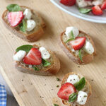 Four slices of baguette topped with fresh basil, mozzarella, and strawberry slices are arranged on a wooden board. More of the ingredients are visible in a bowl in the background. Black pepper is sprinkled on top.