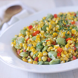 A white plate filled with a colorful vegetable medley of corn, lima beans, and diced red bell peppers, garnished with fresh herbs, sits on a light wooden table. A wooden spoon and cloth napkin are in the background.