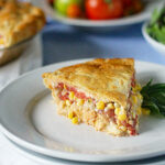 A slice of savory pie filled with corn, tomatoes, and other vegetables sits on a white plate, garnished with a basil leaf. In the background are fresh tomatoes, greens, and the remaining pie.