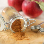 A small glass jar of ground cinnamon is tipped over, spilling onto parchment paper, with metal measuring spoons and three red apples in the background.