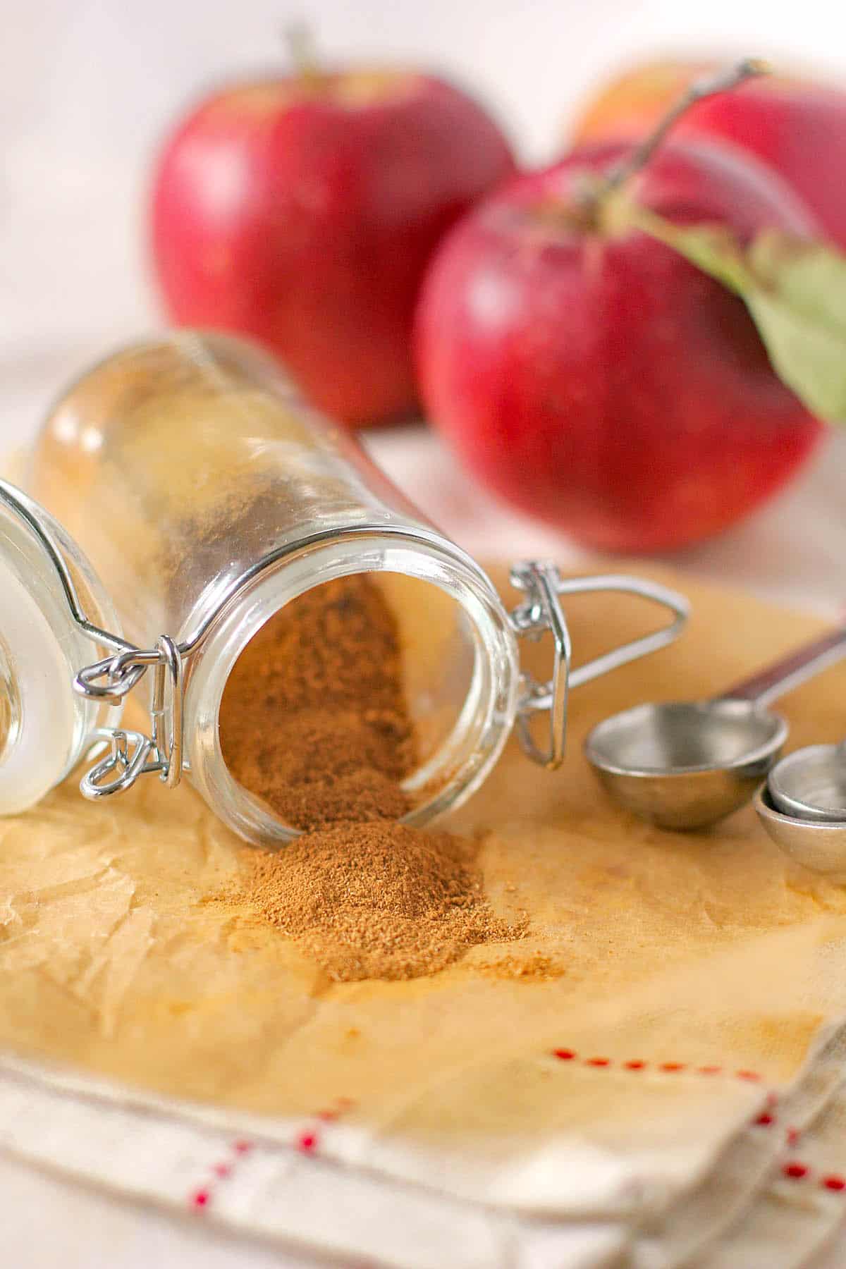 An open glass jar spills homemade apple pie spice onto parchment paper, with stainless steel measuring spoons and red apples in the background.