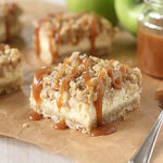 A close-up of cheesecake bars topped with crumbly streusel and drizzled with caramel sauce, set on brown parchment paper with a jar of caramel and a spoon beside them.
