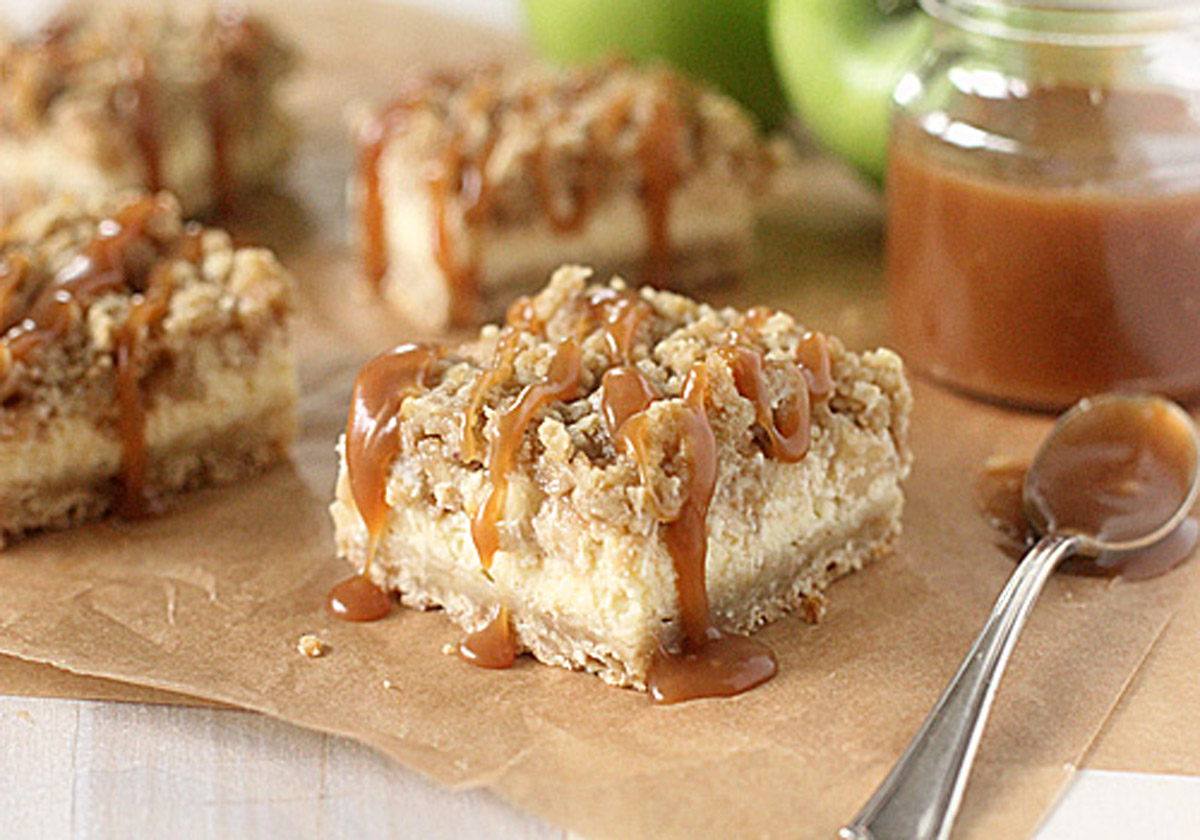 A close-up of cheesecake bars topped with crumbly streusel and drizzled with caramel sauce, set on brown parchment paper with a jar of caramel and a spoon beside them.