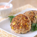 Two golden brown corn and vegetable patties are served on a white plate with fresh basil leaves, next to a small bowl of creamy dipping sauce on a patterned yellow napkin.