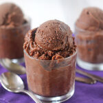 Three clear glasses filled with scoops of rich, dark chocolate sorbet are arranged on a purple cloth, with two metal spoons placed beside them on the table.