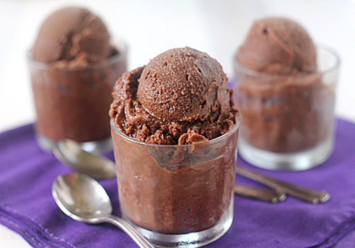 Three clear glasses filled with scoops of rich, dark chocolate sorbet are arranged on a purple cloth, with two metal spoons placed beside them on the table.
