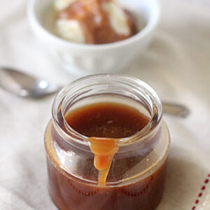 A jar of caramel sauce with some dripping down the side sits on a cloth; in the background, a bowl of ice cream with caramel sauce and a spoon are visible.