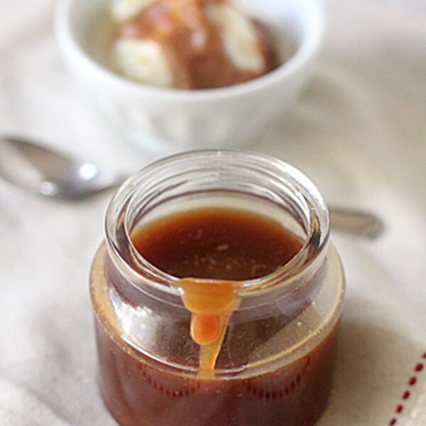 A jar of caramel sauce with some dripping down the side sits on a cloth; in the background, a bowl of ice cream with caramel sauce and a spoon are visible.