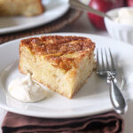 A slice of apple cake sits on a white plate with a dollop of whipped cream and a fork, with another cake, apples, and a bowl in the background.