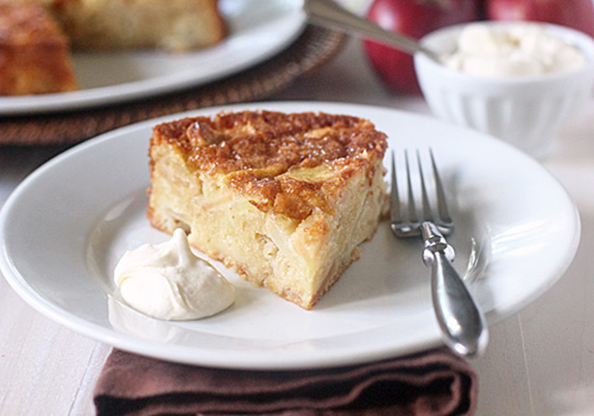 A slice of apple cake sits on a white plate with a dollop of whipped cream and a fork, with another cake, apples, and a bowl in the background.