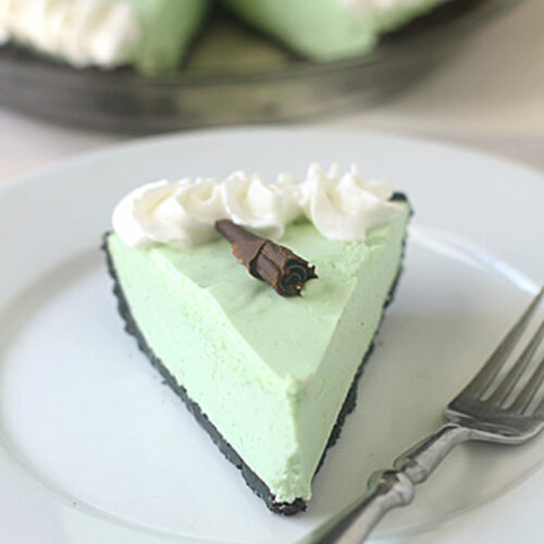 A slice of light green pie with a chocolate cookie crust, topped with whipped cream and a chocolate curl, sits on a white plate next to a fork. The rest of the pie is in the background.