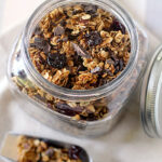 A glass jar filled with homemade granola containing oats, dried fruits, and chocolate chunks sits on a cloth napkin, with a metal scoop of granola beside it. The jar’s metal lid is off to the side.