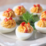 A plate of deviled eggs, each half filled with a yellow creamy mixture and topped with small bits of red pepper, is arranged around a sprig of parsley in the center.