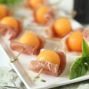 Several skewers of prosciutto-wrapped melon balls are arranged on a white rectangular plate, garnished with fresh basil leaves, set on a light cloth with a drink in the background.