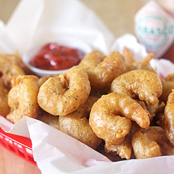 A red basket lined with white paper filled with golden, crispy fried shrimp. A small container of red dipping sauce and a bottle of Tabasco are visible in the background.