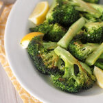 A white plate with roasted broccoli florets and lemon wedges sits on a yellow patterned napkin, with a fork visible in the background.